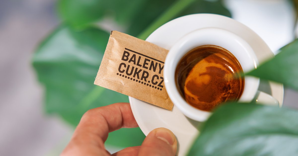 Ruka držící balený cukr s logem nad espresso v bílém šálku